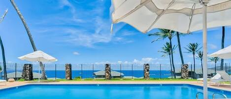 A pool with lounge chairs and umbrellas, a fence with decorative pillars, and a view of the ocean.