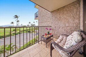 A balcony with a wicker sofa, a small table with a plant and a cup, and a view of a green landscape with palm trees. - *New* Beachfront Kihei Condo near Pickleball and Pool (Kihei)
