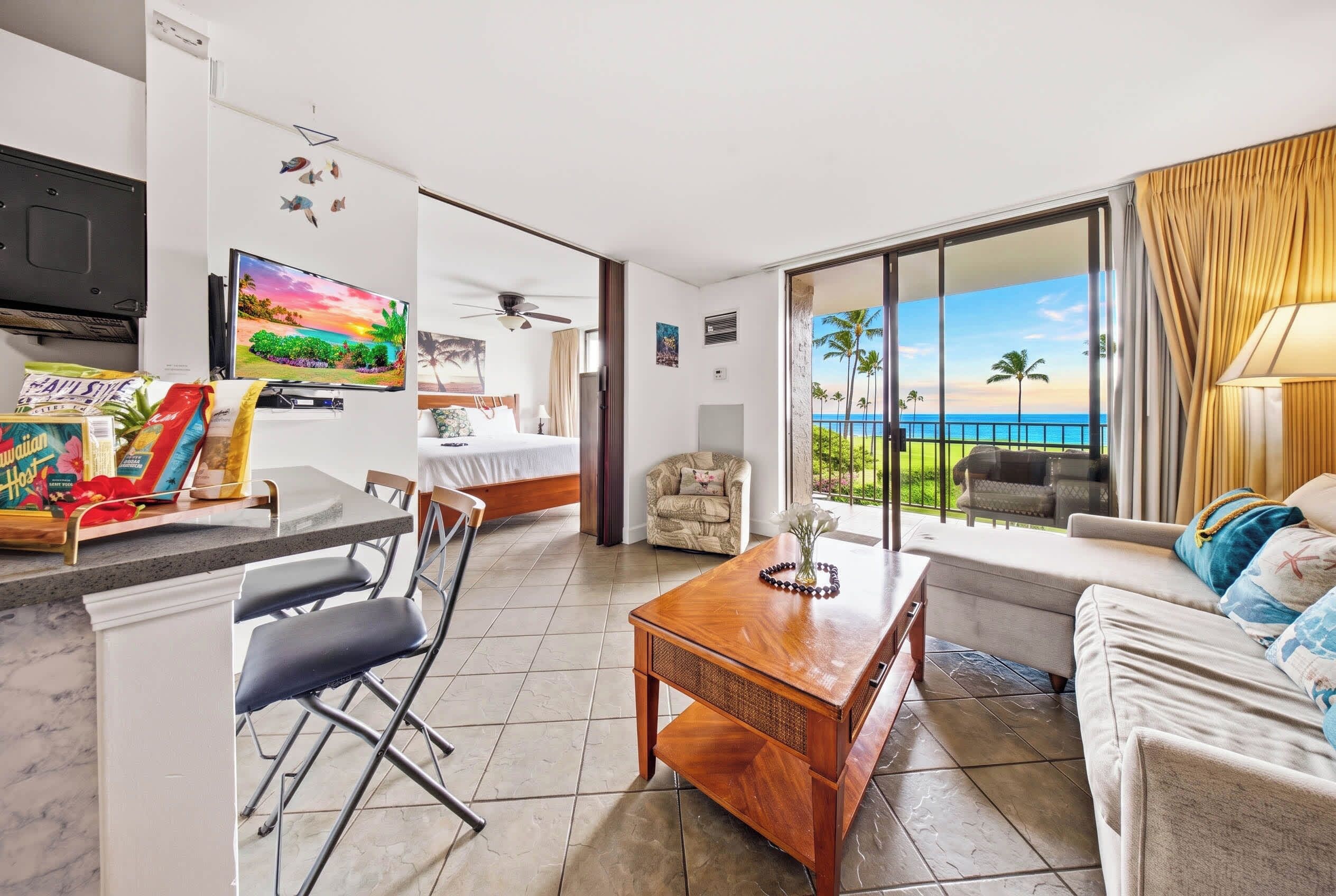A hotel room with a bed, a dining area with a table and chairs, a TV, and a balcony with a view of the ocean.