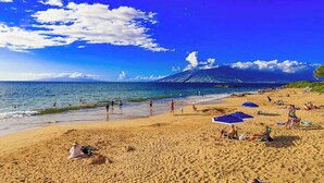 Una playa con gente tomando el sol y nadando, una montaña al fondo y un cielo despejado.