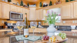 A kitchen with wooden cabinets, a microwave, a wine bottle, glasses, a vase with flowers, and a fruit platter. - Whispering Pines (Sevierville)