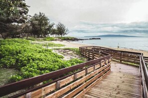 Una pasarela de madera que conduce a una playa con vegetación verde y árboles al fondo.