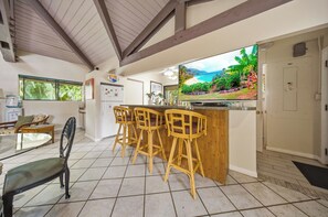 A kitchen with a large TV, wooden bar stools, a dining table with chairs, and a window with a view of greenery. - *New* Fall Deal Oceanfront Kahana Stay, Walk to Pohaku Park (Lahaina)