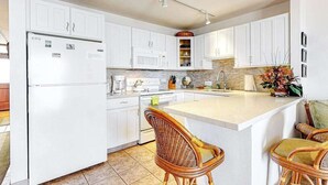 A modern kitchen with white cabinets, a central island, wicker chairs, and a built-in microwave. - New Kihei Ocean-View Retreat for Two with Beachfront Pool (Kihei)