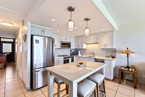 A modern kitchen with stainless steel appliances, a central island with bar stools, pendant lighting, and a view of the adjacent living area. - Ocean-View Kihei Escape for Two, Steps from the Beach (Kihei)