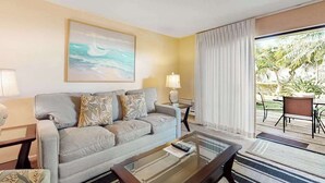 A living room with a sofa, a coffee table, a painting, a lamp, and a view of the outdoors. - NEW Fall Sale Garden-View Kihei 1BR with Pools and Tennis (Kihei)