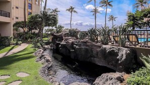 A landscaped area with a rock formation, palm trees, and a pool. - New Beachfront Couple’s Base at Sugar Beach (Kihei)