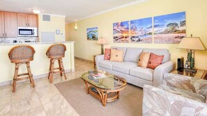 A living room with a sofa, two wicker chairs, a glass coffee table, and a kitchen area with a microwave. - New Beachfront Couple’s Base at Sugar Beach (Kihei)