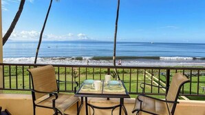 A balcony with two chairs and a table, overlooking a beach with waves and a distant island. - New Beachfront Couple’s Base at Sugar Beach (Kihei)