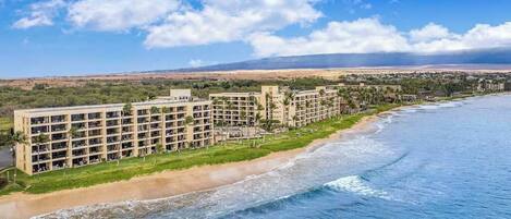 Un hotel frente a la playa con varios pisos, una playa de arena y vistas al océano y colinas distantes.