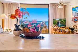 A hotel room with a dining table featuring a tropical fruit bowl, a view of the ocean, and a ceiling fan. - *New* Fall Sale Beachfront Reset with Pool Time at Sugar Beach (Kihei)