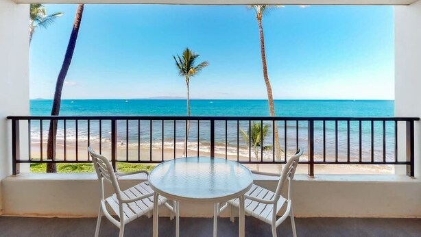 Ein Balkon mit Glastisch und Stühlen, der Blick auf einen Strand mit Palmen und das Meer freigibt.