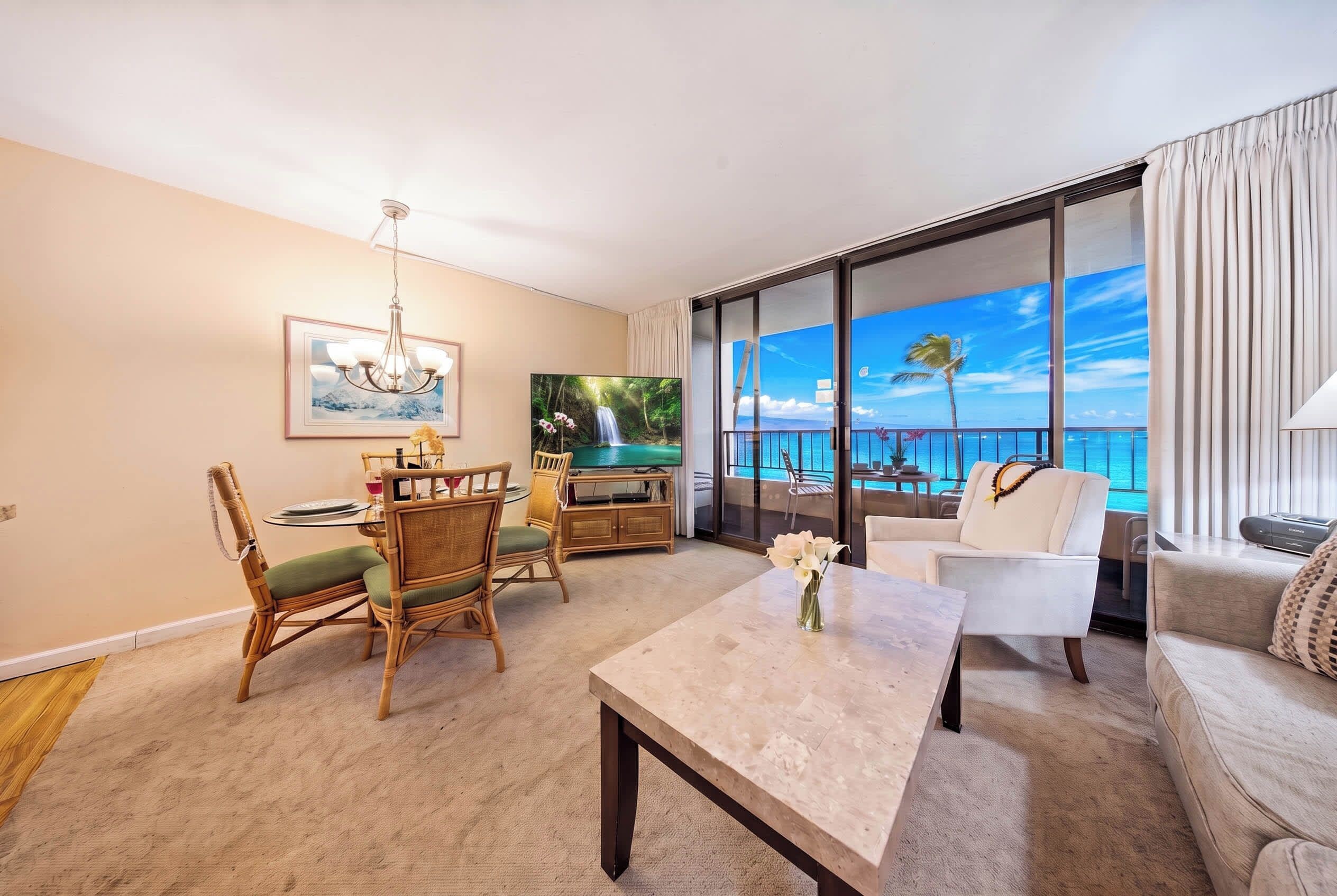A hotel room with a dining area, a living room with a large window overlooking the sea, and a flat-screen TV.