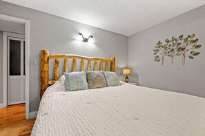 A bedroom with a wooden headboard, a bed with a white textured bedspread, two pillows, a wall-mounted light fixture, and a wall art piece. - Windy Oaks Condo (Gatlinburg)