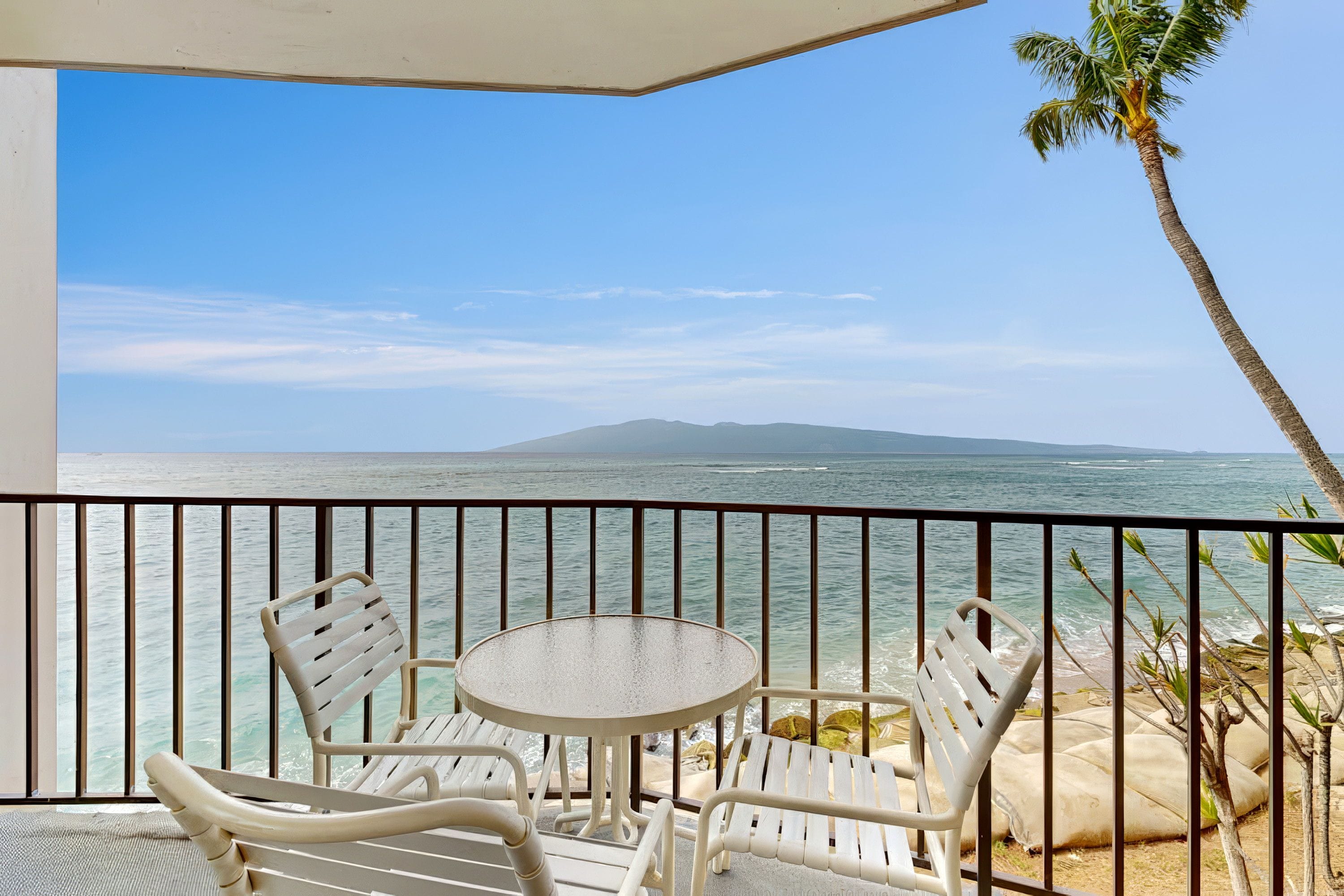 A balcony with a table and two chairs, overlooking a beach with the ocean and a distant island.