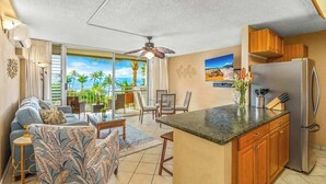 A modern kitchen with a refrigerator, wooden cabinets, and a dining area with chairs and a table. - *NEW* Fall $ale Ocean-View Kihei Escape Steps from The Cove (Kihei)