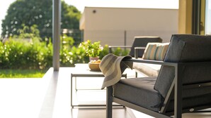 Ein moderner Außenbereich mit Sitzgelegenheiten, darunter eine Couch und ein Sessel, ein kleiner Tisch mit einer Obstschale sowie Blick auf Grünflächen und ein Gebäude im Hintergrund.