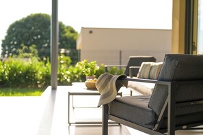 Ein moderner Außenbereich mit Sitzgelegenheiten, darunter eine Couch und ein Sessel, ein kleiner Tisch mit einer Obstschale sowie Blick auf Grünflächen und ein Gebäude im Hintergrund.
