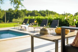 A poolside area with lounge chairs, a table with a hat and fruit bowl, and a view of greenery in the background. - Villa Casa Luta (Istria - Tinjan) (Tinjan)