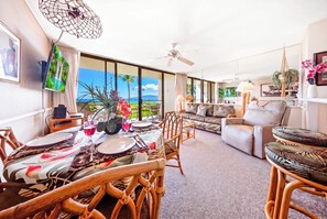 A spacious living area with a large window overlooking a scenic view, a dining area with a table set for two, and a comfortable seating arrangement. - NEW Oceanfront Comfort for Four at Sugar Beach (Kihei)