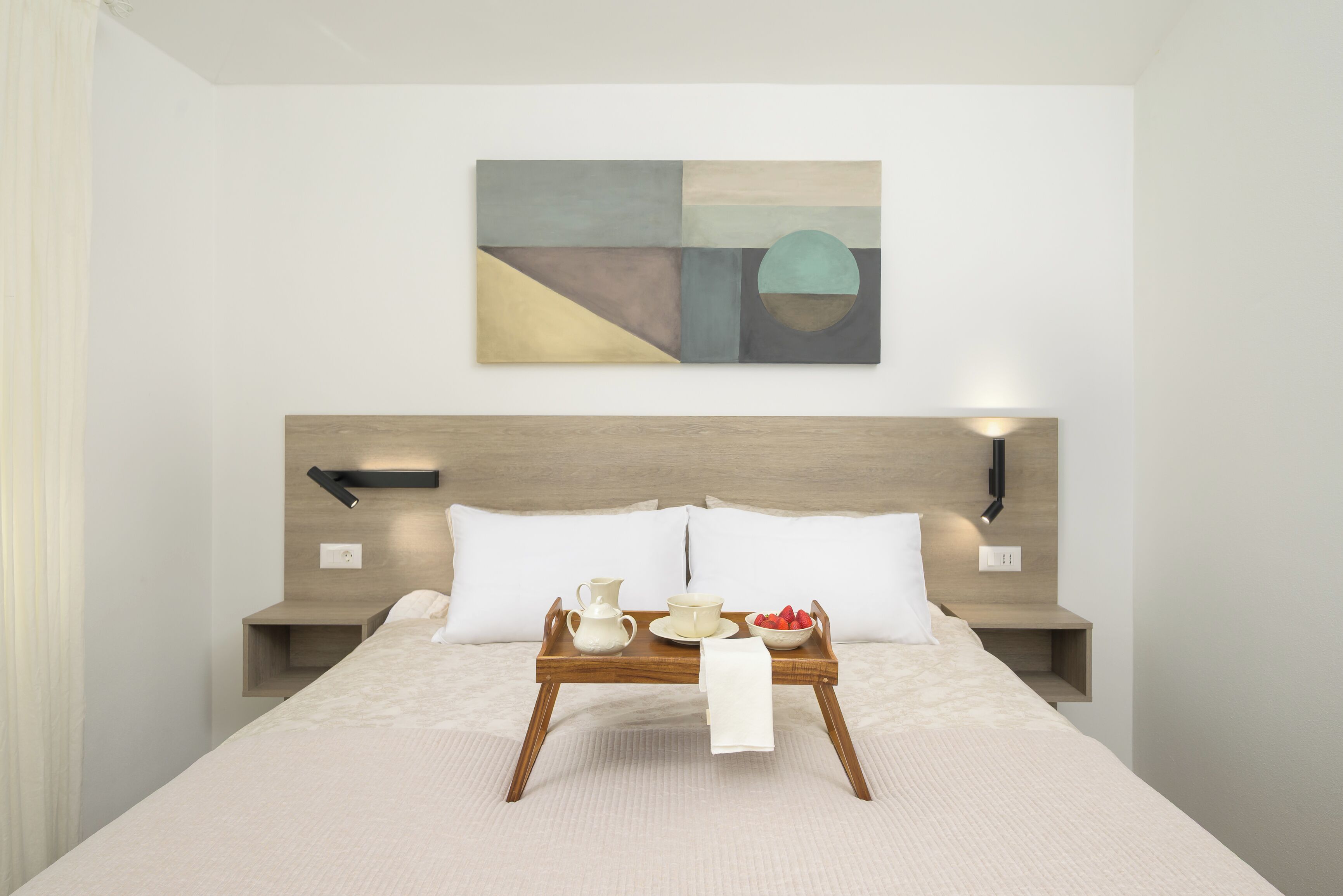 A modern hotel room with a bed, bedside tables, a wall-mounted abstract painting, and a breakfast tray on the bed.