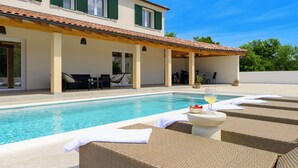 Área de piscina com espreguiçadeiras, uma mesa com um copo de vinho e vista para uma casa com persianas verdes.