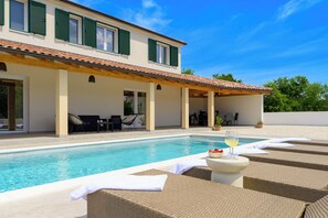 Ein Poolbereich mit Liegestühlen, einem Tisch mit einem Weinglas und Blick auf ein Haus mit grünen Fensterläden.