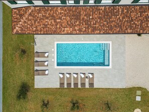Eine Vogelperspektive auf ein rechteckiges Schwimmbecken mit einer gefliesten Liegefläche, umgeben von Liegestühlen und einem überdachten Bereich mit einem Flachdach.