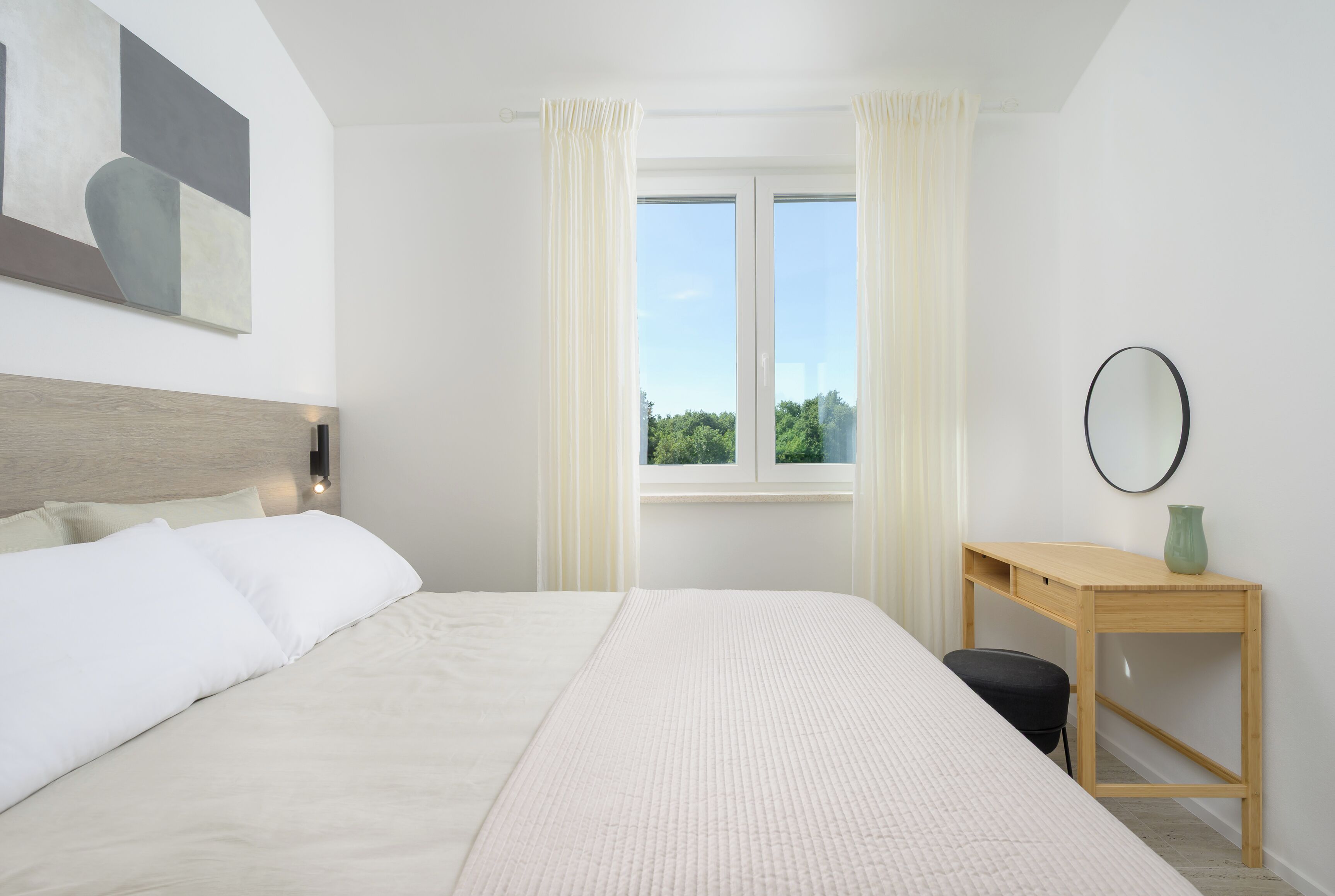 A bedroom with a bed, a wooden desk, a window with curtains, and a wall-mounted mirror.