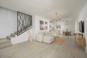 Un salon moderne avec un canapé d’angle, un coin repas avec une table et des chaises, un escalier agrémenté d’une œuvre d’art murale aux motifs géométriques et un lustre.