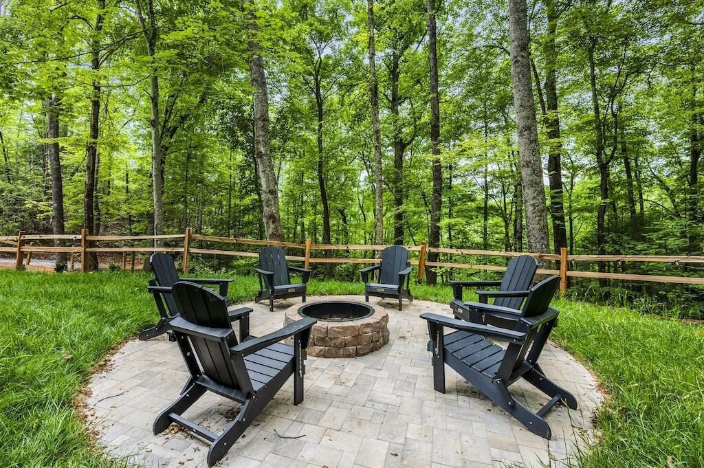 An outdoor seating area with Adirondack chairs and a stone fire pit surrounded by a wooden fence, set in a forested area.