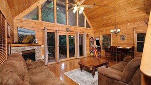 A spacious living room with a stone fireplace, large windows, and a ceiling fan. - Yeti Lodge (Gatlinburg)