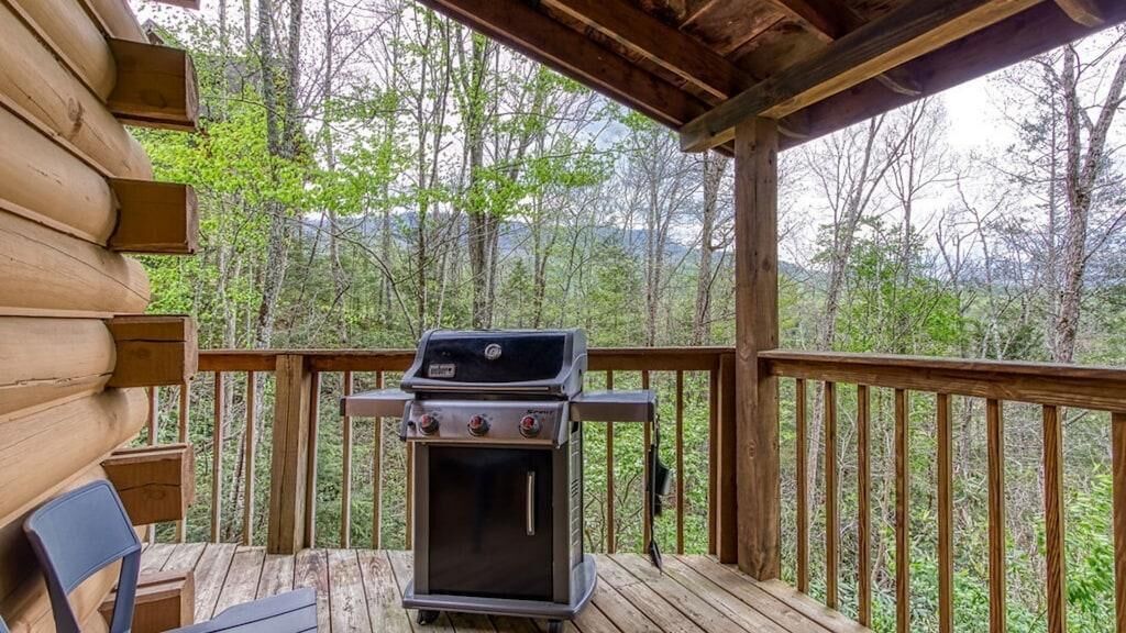 Terraza de madera con una parrilla y sillas, con vista a un área boscosa.