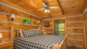 A log cabin bedroom with a bed, a nightstand, a ceiling fan, and a window with blinds. - Tiny Pines (Sevierville)