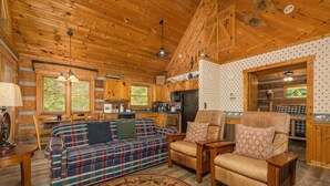 A log cabin interior with a plaid sofa, leather armchairs, a dining table, and a kitchen area. - Tiny Pines (Sevierville)