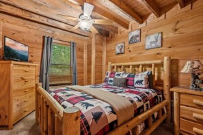 A wooden cabin bedroom with a bed, a dresser, a nightstand, a ceiling fan, and a window with blinds. - Friends in High Places 2 (Sevierville)