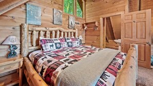 Dormitorio de cabaña de madera con cama, mesita de noche, lámpara y ventana con vista a los árboles.
