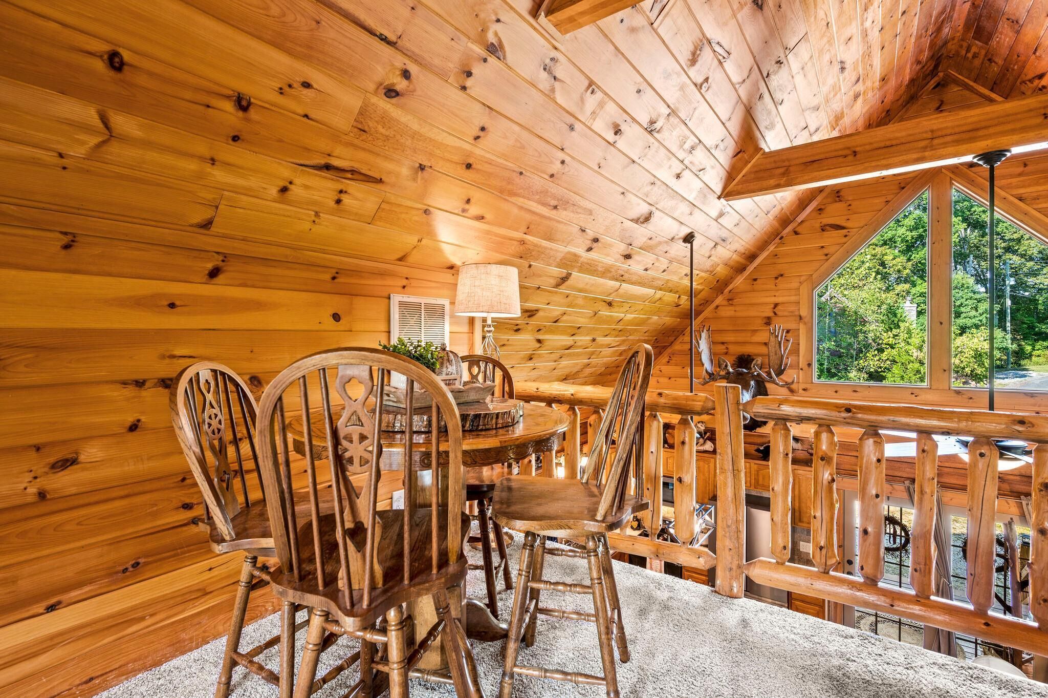 A wooden interior with a dining table and chairs, a lamp, and a window with a view of trees.