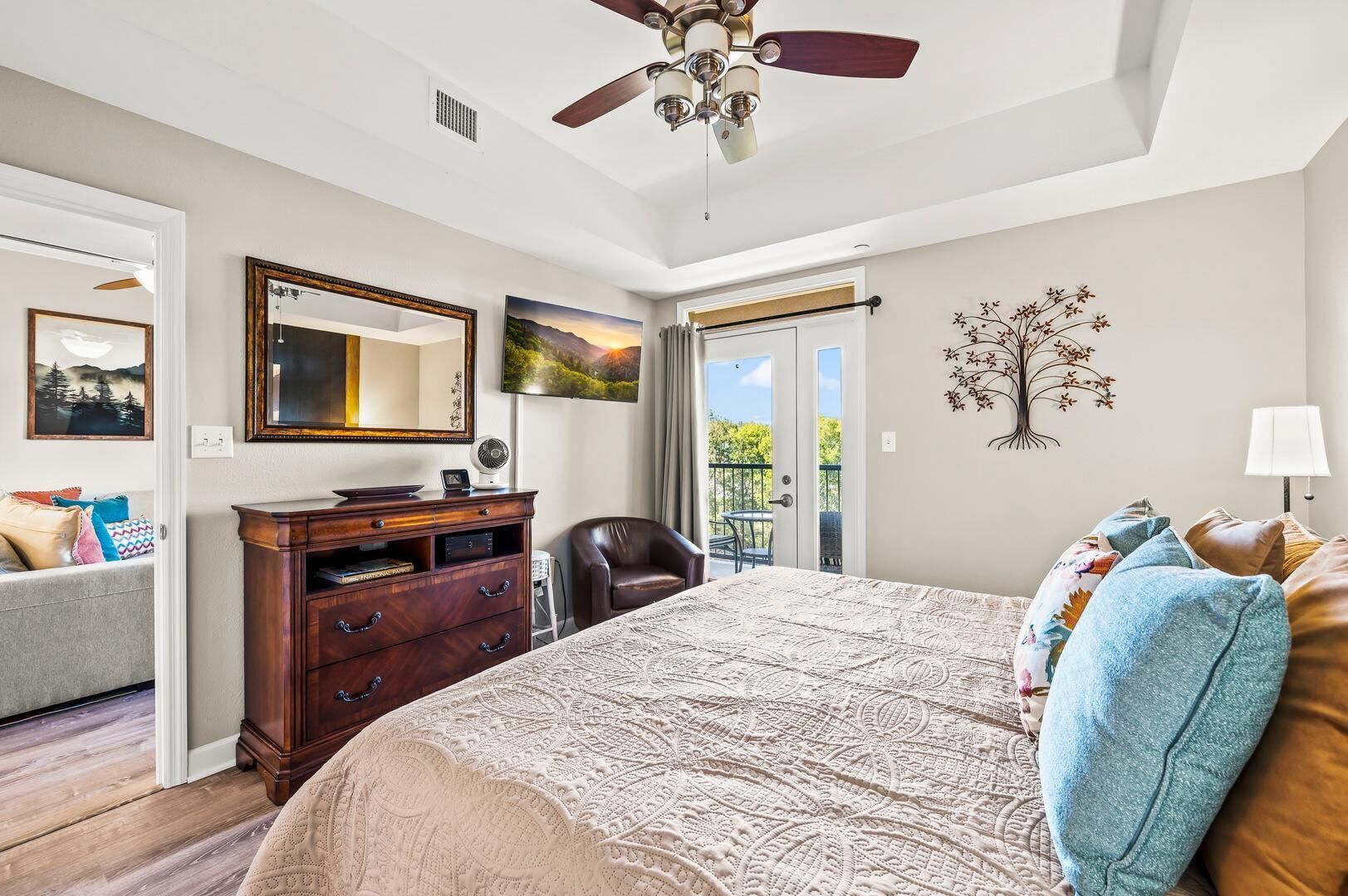 A bedroom with a large bed, a dresser, a chair, a ceiling fan, a mirror, a TV, and a balcony door.