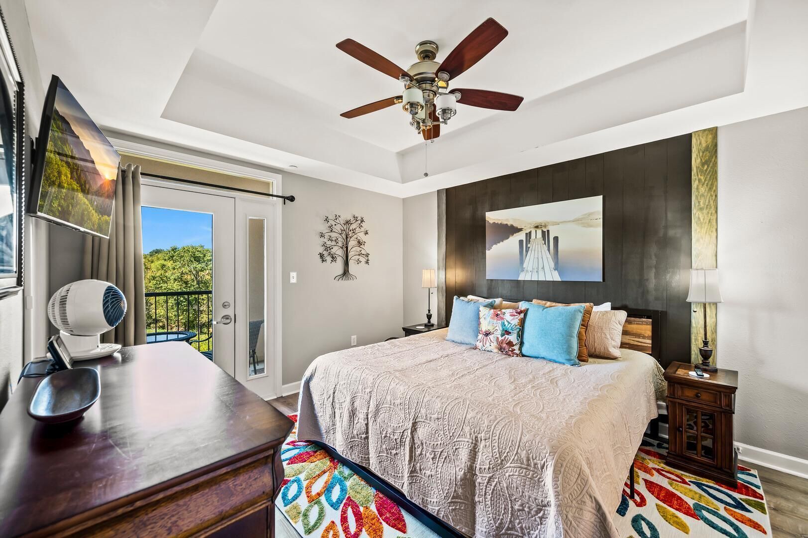 Une chambre avec un lit, un ventilateur de plafond, un bureau avec une chaise, une fenêtre avec des rideaux et une vue sur la verdure à l’extérieur.