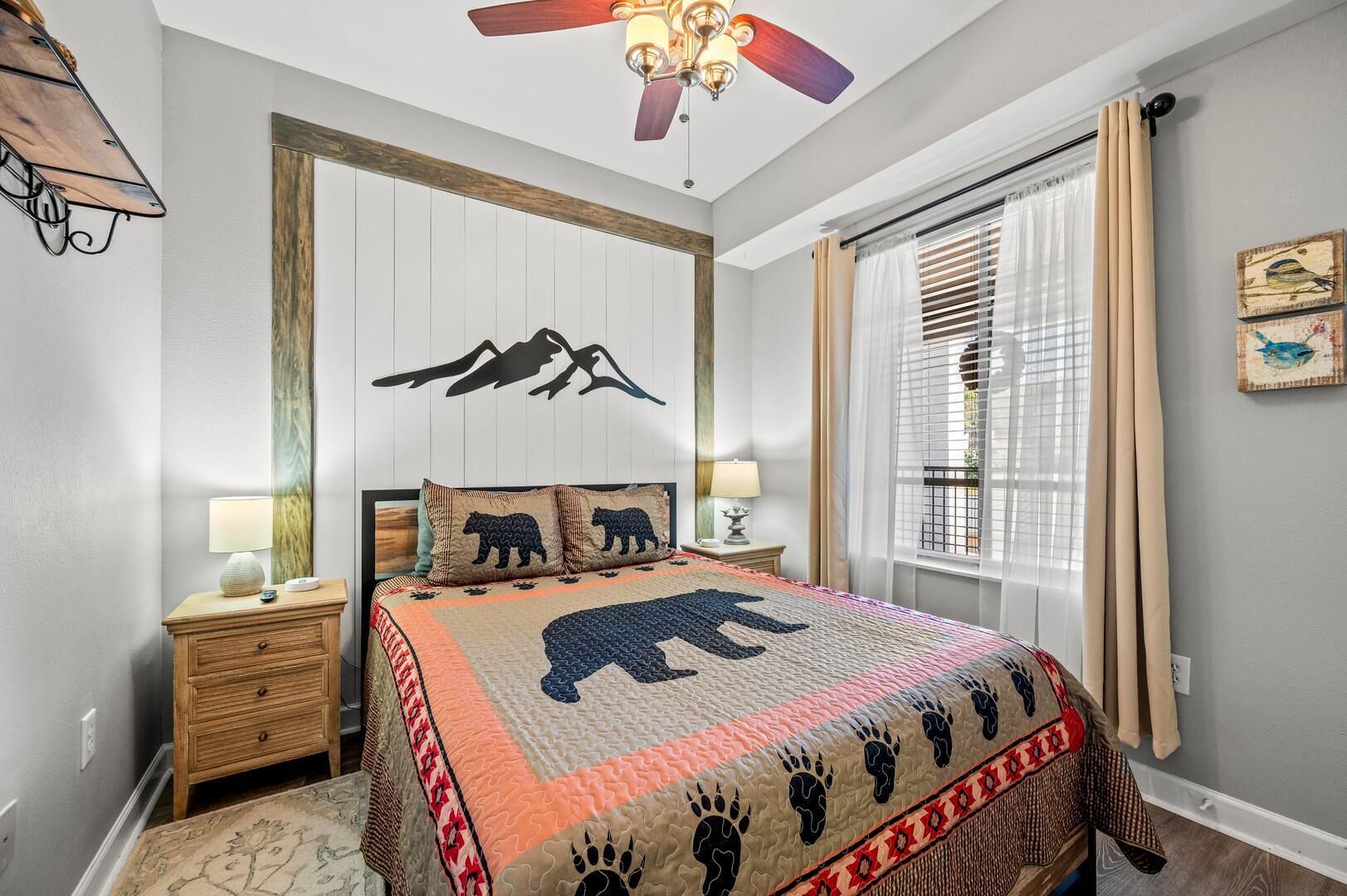 A bedroom with a bed, a nightstand, a lamp, a ceiling fan, a window with curtains, and wall art.