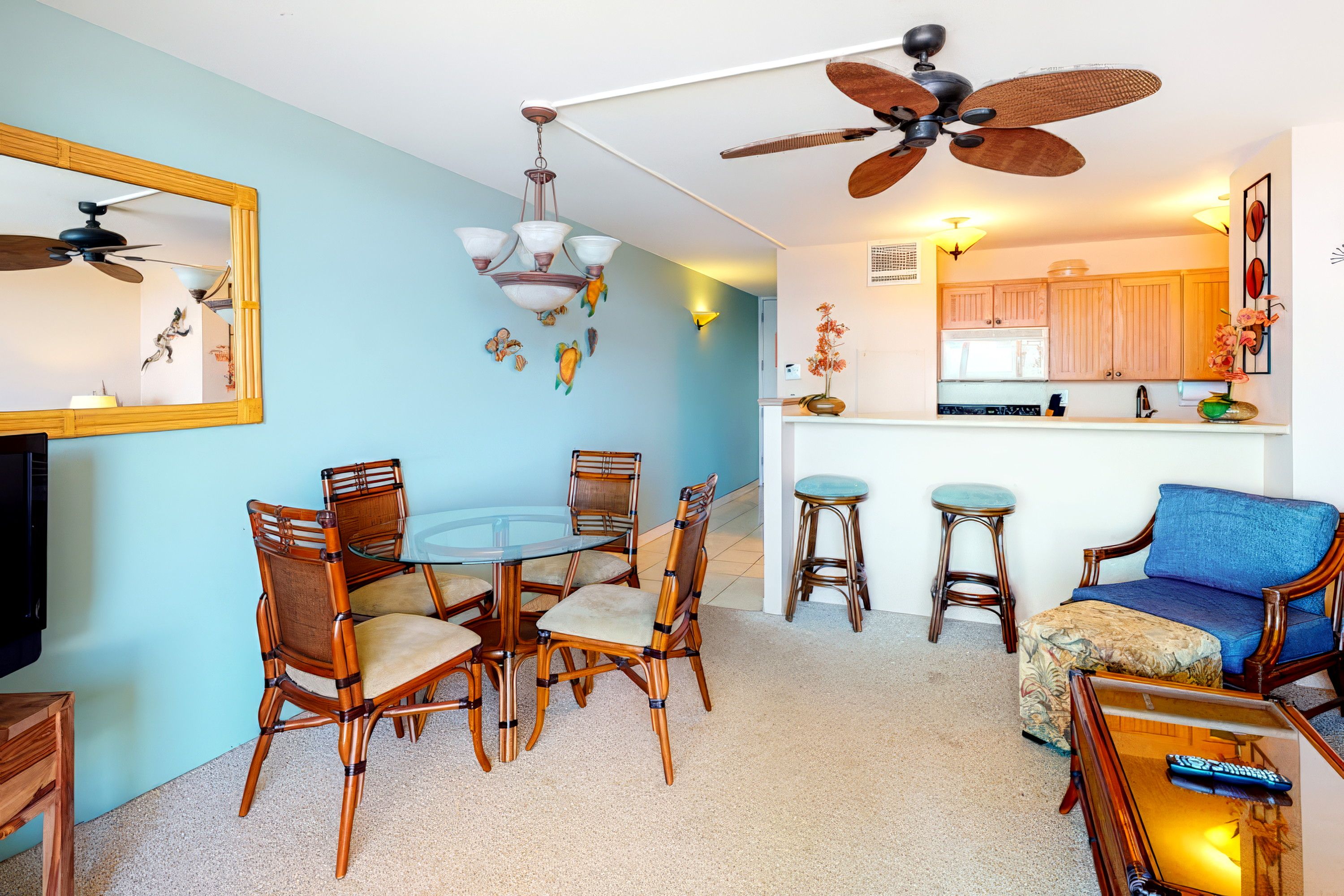 A dining area with a round glass table and chairs, a ceiling fan, and a kitchen area with wooden cabinets and a bar stool.