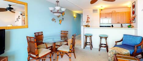 A dining area with a round glass table and chairs, a ceiling fan, and a kitchen area with wooden cabinets and a bar stool.