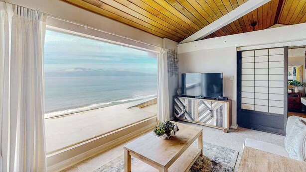 Ein Wohnzimmer mit großem Fenster zum Meer, einem Flachbildfernseher und einer Holzzieldecke.