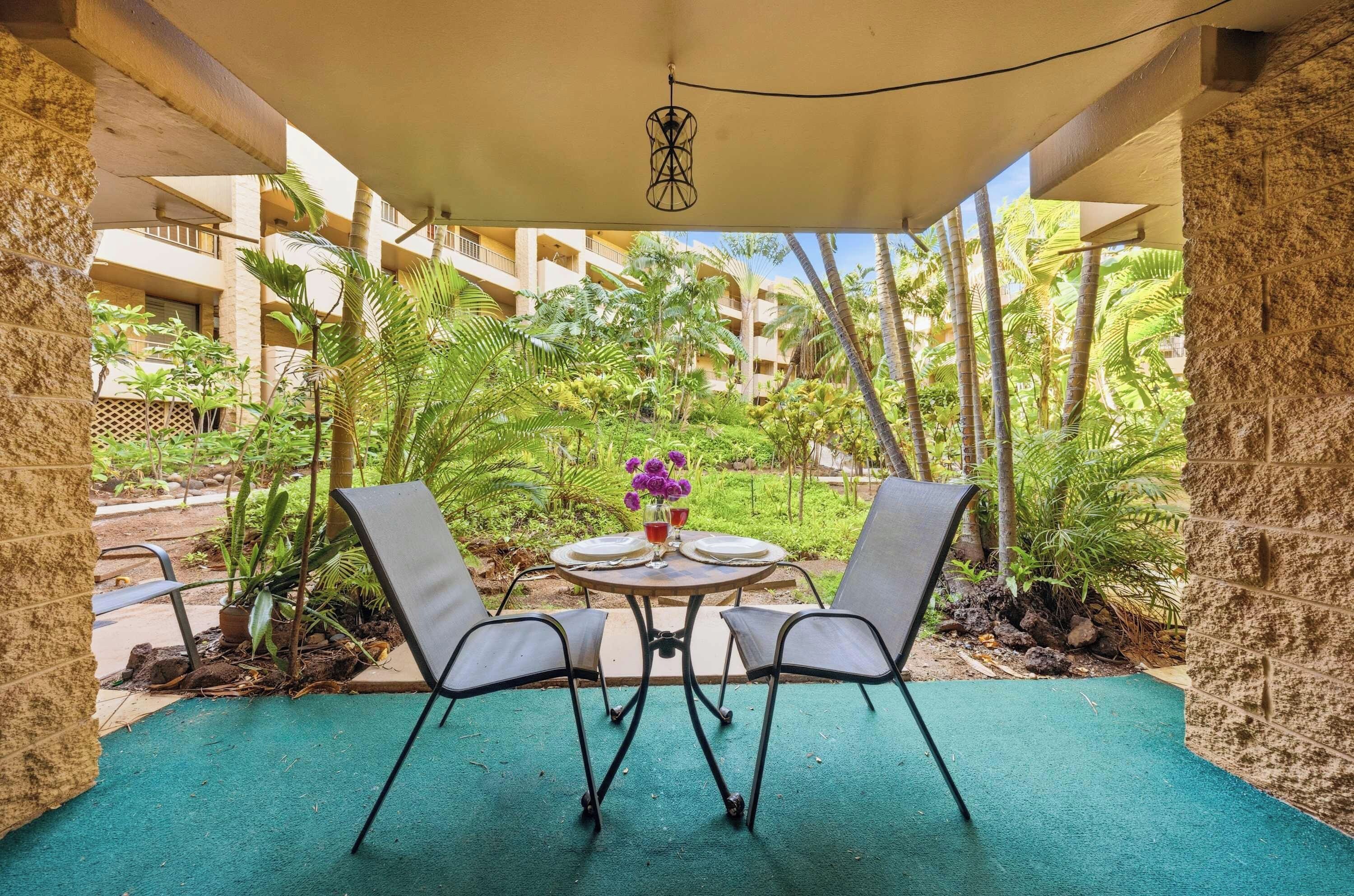 A covered outdoor seating area with two chairs and a table, surrounded by lush greenery and a view of a building in the background.