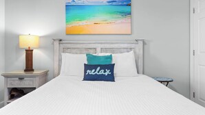 A bed with a white quilt, a teal pillow with 'relax' text, a wooden nightstand with a lamp, and a painting of a beach. - Majestic View Oceanfront (Panama City Beach)