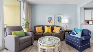 A living room with a grey armchair, a brown sofa with yellow cushions, a blue sofa with a whale pillow, a round glass table, and a potted plant. - Majestic View Oceanfront (Panama City Beach)
