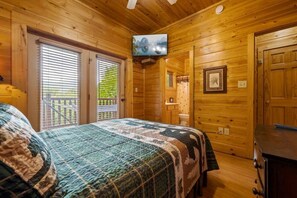 A wooden cabin bedroom with a bed, a television mounted on the wall, and a door leading to an outdoor area. - Mountain Sunrise (Sevierville)