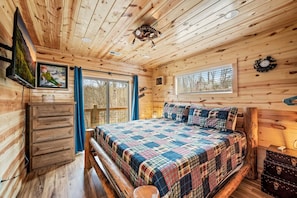 A wooden cabin interior with a bed, a mounted television, and a window with a view of trees. - Neverland (Gatlinburg)