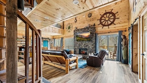 A wooden cabin interior with a stone fireplace, a sofa, a chair, and a staircase. - Neverland (Gatlinburg)